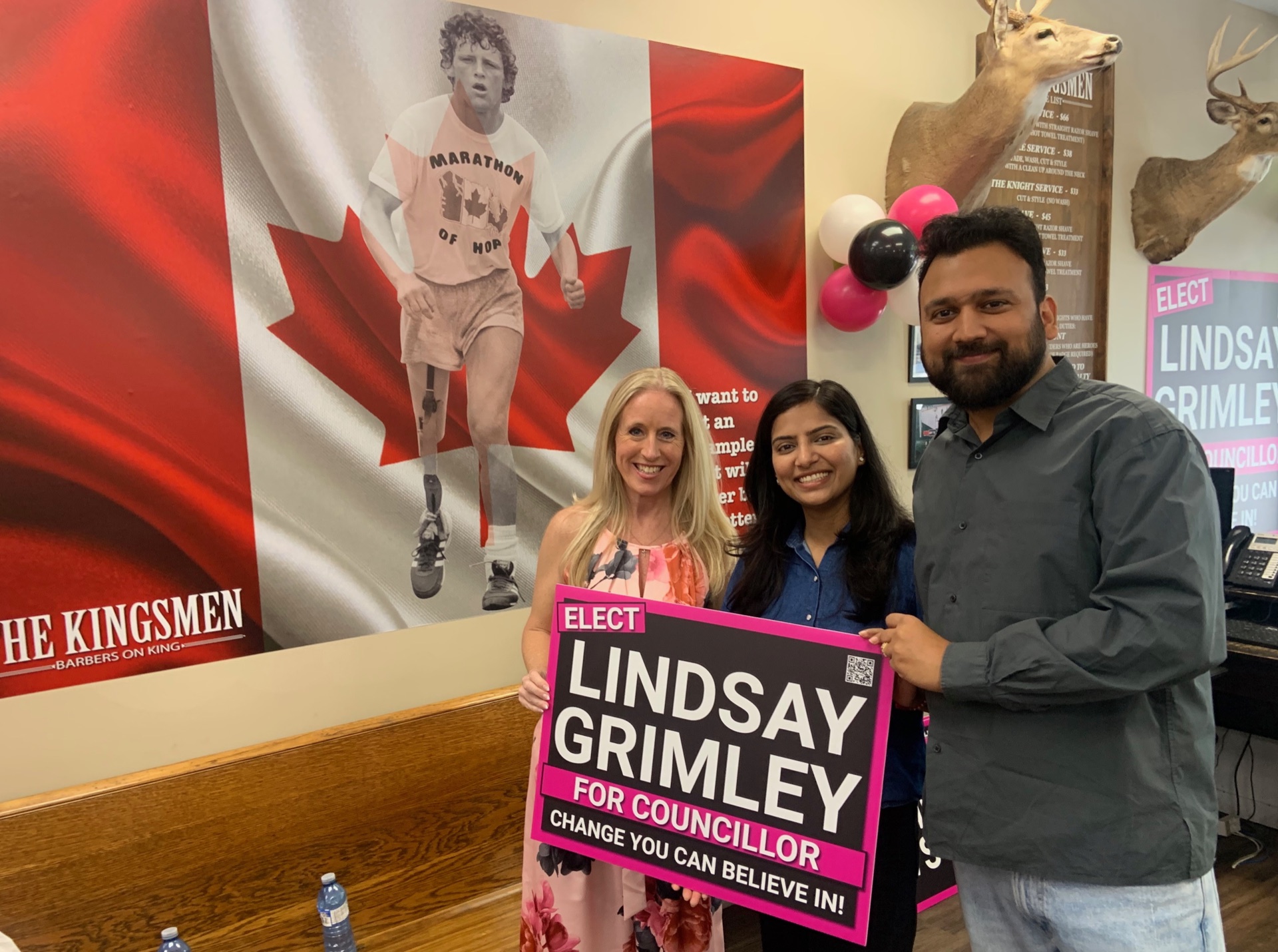 Lindsay Grimley with two supporters holding a campaign sign in front of the Terry Fox mural at The Kingsmen Barbers on King