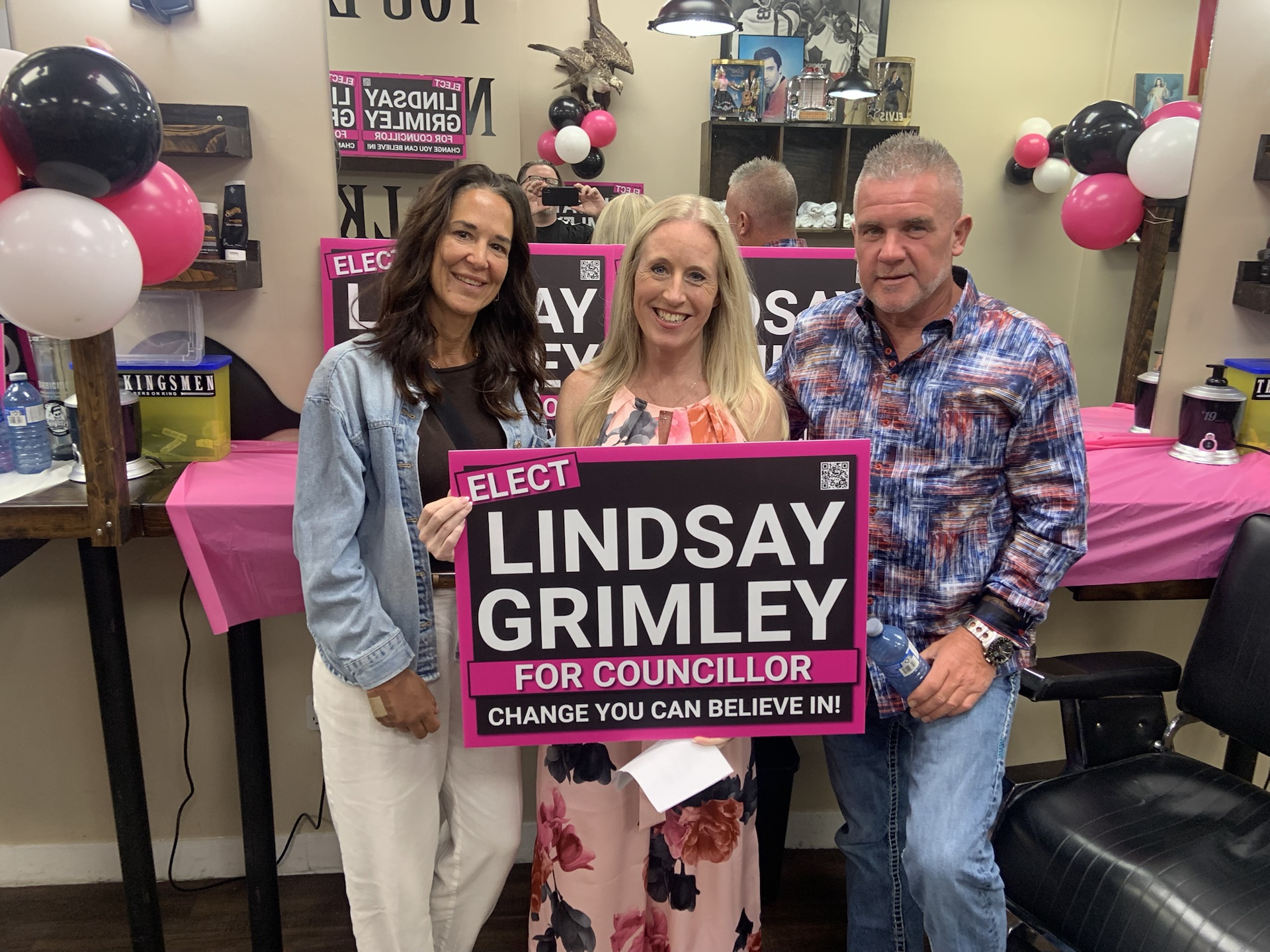 Lindsay Grimley with two supporters holding a campaign sign in the barber shop during the launch party