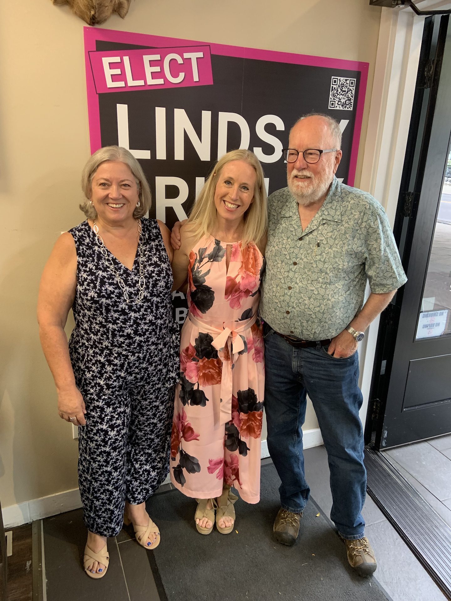 Lindsay Grimley with two guests in front of an Elect Lindsay Grimley campaign poster