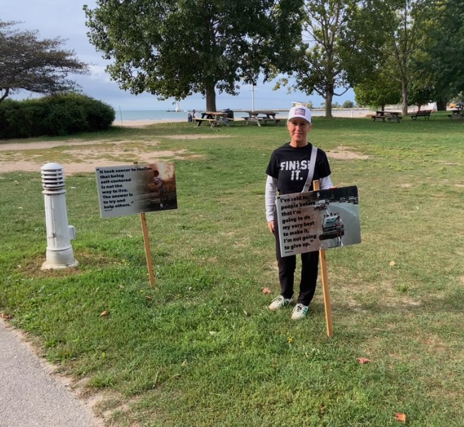 Lindsay Grimley in a Finish It shirt stands in Victoria Park by the water, with two Terry Fox signs planted in the grass.
